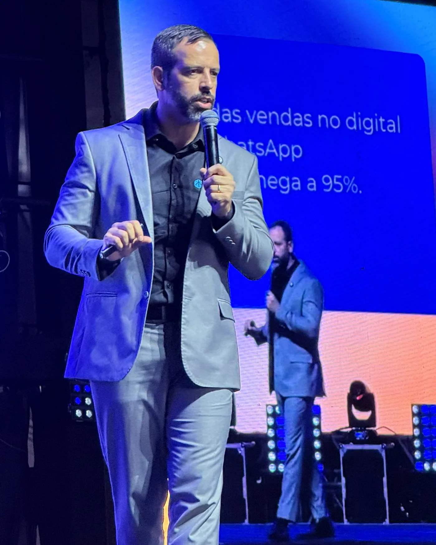 Rafael Tonietto palestrando em um palco, de blazer, com telão ao fundo