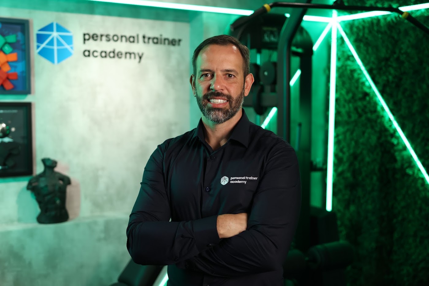 Rafael Tonietto de braços cruzados, de camisa preta com o logo da Personal Trainer Academy, em estúdio com luz verde