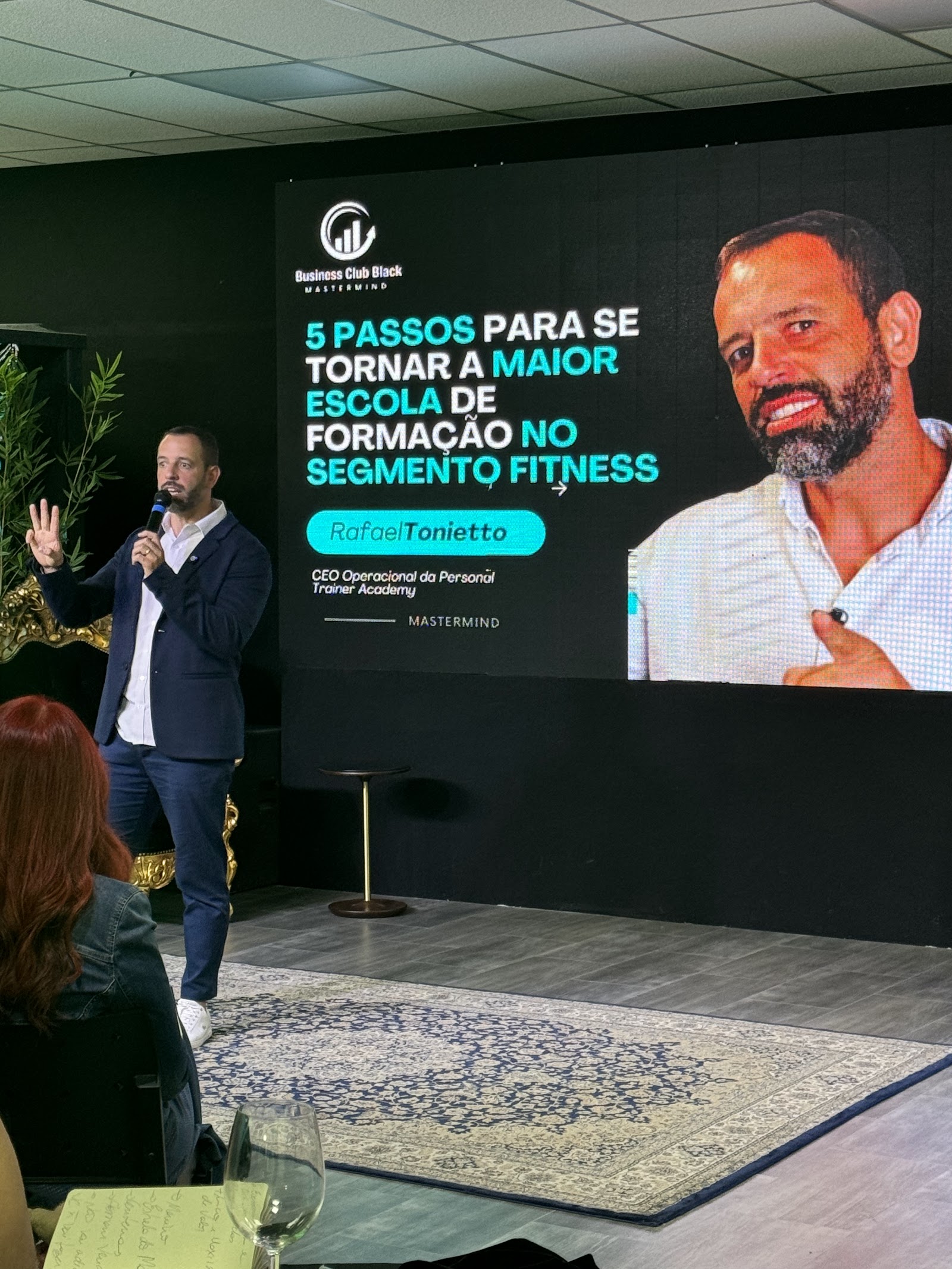 Rafael Tonietto de blazer falando ao microfone num palco, com o telão do Business Club Black Mastermind ao fundo